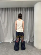 Pantalón de jean azul raw con diseño de corte versátil, ajustable mediante cierres laterales que revelan un panel interior rojo. Presenta una estampa gráfica de gran tamaño en la parte trasera con detalles bordados, costuras reforzadas y etiqueta de cuero en la cintura.