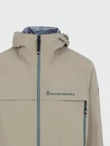 Campera impermeable Moncler modelo Grenoble Shipton en color beige, con capucha ajustable, cierre frontal de cremallera y bolsillos laterales con cierre. Presenta el logo de la marca estampado en el pecho.