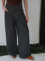 Pantalón wide leg de tiro medio confeccionado en denim color gris oscuro. Presenta una cintura trabajada con detalles de pasacintas entrelazadas, cierre frontal y bolsillos laterales. Su diseño de pierna amplia aporta volumen y movimiento.