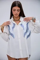 Camisa blanca de manga larga con cuello clásico y botones frontales. Presenta un diseño de ondas en tonos azules con lentejuelas y pequeños apliques metálicos en el frente.