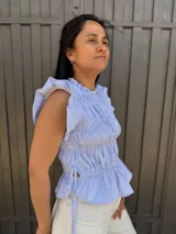 Blusa sin mangas a rayas finas en celeste y blanco, con cuello redondo alto y volados en los hombros. Presenta frunces en el frente, lazos laterales regulables y bajo con ligero vuelo.