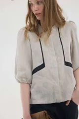 Camisa de lino color beige, de silueta romántica con escote a la base y mini volado. Cuenta con mangas 3/4 abullonadas con frunces y terminación en elástico. Posee detalles de puntillas negras en contraste en el frente y cierre frontal con botones ocultos bajo cartera.