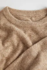 Sweater de punto de textura suave y diseño holgado, con cuello redondo y mangas largas. Presenta un tono beige melange y un corte ligeramente más corto en la parte frontal.