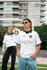 Camiseta de fútbol estilo boxy color blanco, con cuello polo y ribetes negros en las mangas. Presenta detalles estampados y bordados exclusivos en el frente y hombros.