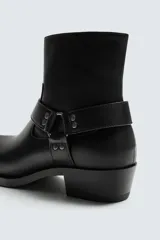 Bota de media caña en color negro, con diseño de arnés que incluye tiras, argollas metálicas y tachas decorativas alrededor del tobillo. Presenta punta fina y tacón cuadrado.