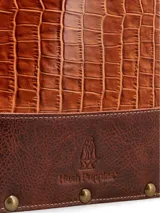 Bolso matero Hush Puppies de cuero en tono tostado con textura símil cocodrilo y detalles en marrón oscuro en la base. Cuenta con doble asa larga para hombro y tachas metálicas decorativas.