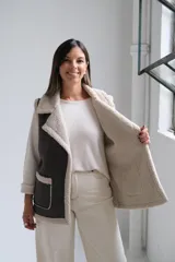 Chaleco de gamuza sintética en color gris oscuro con interior y bordes de corderito beige. Presenta solapas amplias, dos bolsillos frontales con ribetes de corderito y diseño sin cierre.