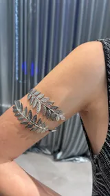 Brazalete de brazo abierto en color plateado, con un diseño envolvente que presenta dos ramas con hojas detalladas en los extremos.