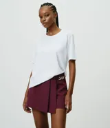 Short pollera de sastrería marca Cortelle, color bordó, confeccionado en crepé. Presenta sobreposición frontal efecto pollera con pliegues y detalle de cadena metálica decorativa en el lateral.