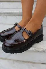 Zapato tipo guillermina en cuero charol color bordó, con suela track negra de goma, cierre con hebilla ajustable y detalle de herrajes metálicos dorados.