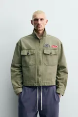 Cazadora cropped de algodón con efecto lavado, color verde militar. Presenta cuello solapa, cierre frontal con cremallera, bolsillos frontales de plastrón con solapa y parches decorativos en el pecho.