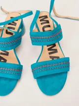 Sandalias de tacón bajo Mutma modelo Cunaza, color turquesa, confeccionadas en gamuza con detalles de costuras y bordados en hilo naranja. Tienen tiras dobles en el empeine y una tira fina ajustable alrededor del tobillo.