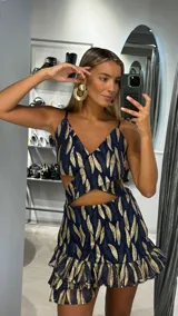 Vestido corto con diseño cut-out en la cintura, tirantes finos y falda con volados en el ruedo. Presenta un estampado de hojas en tonos azul marino y dorado con acabado brillante.