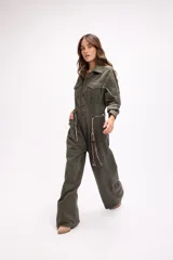 Mono de estilo utilitario color beige, con cierre frontal de botones, bolsillos en el pecho y cintura entallada con detalle de cadenas decorativas. Presenta mangas largas y piernas de corte recto y holgado.