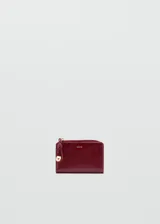 Billetera pequeña de diseño compacto en acabado charol color bordó. Cuenta con cierre perimetral con tirador decorativo de cordón y detalle metálico dorado, y logo de la marca grabado en el frente.