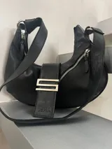Cartera tipo hobo con diseño de media luna, confeccionada en cuero negro. Presenta una solapa frontal con un detalle metálico en forma de letra S y el logo de la marca grabado. Cuenta con cierre superior y correa ajustable para llevar al hombro.