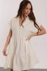 Vestido corto color crudo con cuello camisero y escote en V. Presenta cierre frontal parcial con botones, detalles de puntilla vertical en el pecho y falda con volado en el ruedo.