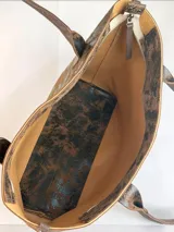 Cartera tipo tote de gran tamaño, confeccionada en cuero genuino con acabado texturizado y efecto desgastado en tonos tierra y negro. Cuenta con cierre superior metálico, interior forrado en cuero y un sujetador de asas con broche decorativo.