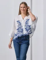 Blusa blanca con bordado floral azul, escote en V, manga japonesa y cierre frontal con botones.