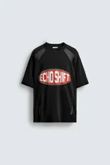 Camiseta negra de corte boxy fit con paneles laterales y de hombros en tejido de malla. Presenta un estampado frontal ovalado con borde rojo y texto blanco que dice "ECHO SHIFT".