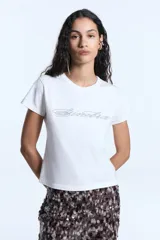 Remera de jersey de algodón, de calce ajustado, con escote redondo y manga corta. Presenta una estampa frontal con diseño de letras en glitter.
