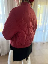Campera tipo bomber acolchada con diseño de pespuntes ondulados, cierre frontal y bolsillos laterales con botón. Presenta un corte moderno y acabado en color bordo.