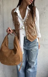 Chaleco de gamuza color camel con bordados en hilo blanco y detalles de tachas metálicas. Presenta un diseño abierto con terminaciones redondeadas en el frente.