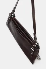 Bolso de hombro de formato alargado, color marrón oscuro con acabado ligeramente desgastado. El asa de hombro es delgada y ajustable mediante hebillas metálicas. Presenta detalles de tachas o remaches metálicos en los puntos de unión del asa.