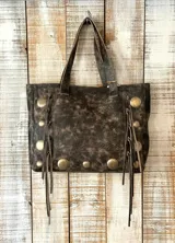 Cartera estilo shopper de cuero con acabado desgastado, decorada con tachas metálicas circulares y flecos laterales. Cuenta con doble asa superior.