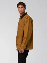 Chaqueta tipo chore de lona color marrón, con cuello camisero en contraste color marrón oscuro, cierre frontal con botones, cuatro bolsillos de parche frontales y costuras a la vista en color blanco.
