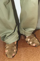 Sandalia plana tipo cangrejera en serraje color marrón claro, con tiras entrelazadas en la parte delantera y cierre de hebilla en el tobillo. Tiene punta redonda y un tacón bajo de 2 cm.