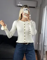 Saquito de tejido bremer color crema, con cuello redondo, cierre frontal de botones y detalles de volados en el ruedo y los puños.