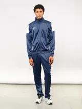 Conjunto deportivo Umbro compuesto por campera de cuello alto con cierre frontal y pantalón de corte recto. La campera presenta bloques de color blanco en los hombros y el logo de la marca bordado en el pecho. Confeccionado en tejido sintético con acabado satinado.