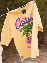 Remera color amarillo pastel con estampado de palmeras, flores de hibisco y texto 'California Ride the Tide Rusty Tribe'.