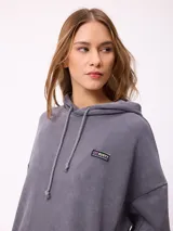 Canguro oversize de color gris con efecto lavado, capucha con cordones ajustables, bolsillo frontal tipo canguro y parche pequeño con logo de la marca en el pecho.