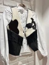 Chaleco corto confeccionado en cuero vacuno con pelo en la parte superior con estampado de vaca en blanco y negro, y cuero liso negro en la parte inferior con flecos colgantes.