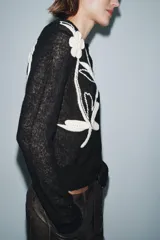 Chaqueta de punto negra con cuello redondo y manga larga. Presenta bordados florales en color blanco en la parte delantera y cierre frontal con botones.