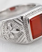 Anillo de acero tipo sello con piedra central rectangular de color rojo y diseño grabado en los laterales.