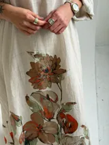 Vestido largo beige con diseño holgado, escote en V y mangas largas, adornado con un gran estampado floral en tonos marrones y verdes en la parte inferior.