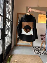 Campera tipo hoodie de textura tipo corderito en color negro, con diseño de collage en la espalda que combina parches en tonos marrón, blanco y estampado animal print de leopardo.