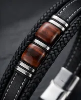 Pulsera de diseño masculino compuesta por una banda de cuero sintético trenzado en color negro, con detalles centrales de acero y piedras naturales ojo de tigre.