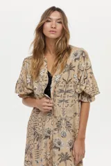 Vestido túnica de lino y viscosa, color camel con estampa artesanal en tonos oscuros, escote en V profundo, mangas amplias abullonadas y falda larga con volado en el ruedo.