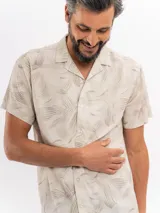 Camisa de manga corta color beige con estampado tropical de hojas, cuello camisero abierto y cierre frontal de botones.