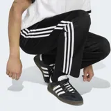 Pantalón deportivo Adidas Firebird de corte recto y holgado, color negro con las clásicas tres tiras blancas a lo largo de los laterales y el logo del trifolio bordado en la pierna izquierda.