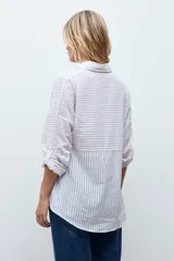 Camisa de corte holgado con diseño de rayas verticales en tonos beige y blanco, con sutiles destellos brillantes. Presenta cuello clásico, cierre frontal con botones y mangas largas con puños ajustables.