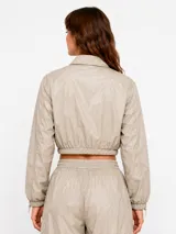 Campera corta de nylon color beige, con cuello camisero, cierre frontal con botones a presión, mangas largas con puños elásticos y cordón ajustable en la cintura.