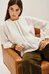 Camisa de algodón blanco con textura de pequeños puntos en relieve, cuello redondo con cierre de botones oculto, detalles de alforzas verticales y encaje en el frente, y mangas largas con puños elásticos.