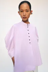 Camisa confeccionada en hilatura de algodón de color rosado claro, con cuello mao y cierre frontal parcial con botones a contraste. Presenta mangas por debajo del codo abullonadas y bajo asimétrico con aberturas laterales.