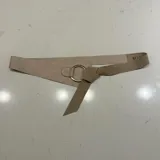 Cinto de gamuza color beige con diseño asimétrico, cuenta con un aro metálico dorado en el centro y un nudo decorativo.