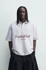 Camisa polo boxy fit de manga corta, confeccionada en tejido de algodón con diseño de rayas verticales celestes y blancas. Cuenta con cuello solapa, cierre frontal con botones y estampado frontal de texto en tono marrón con la leyenda "Poetic Work Since 1975".